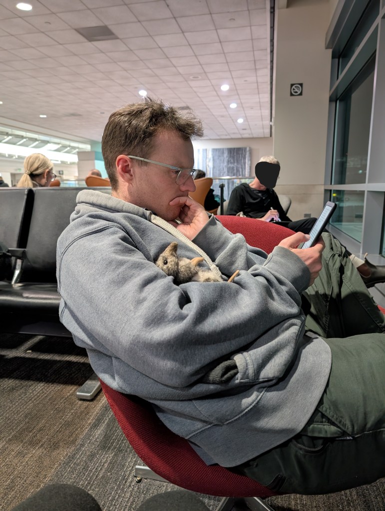 a man sits in an airport and looks at a phone. in the crook of his arm sits a stuffed animal rat