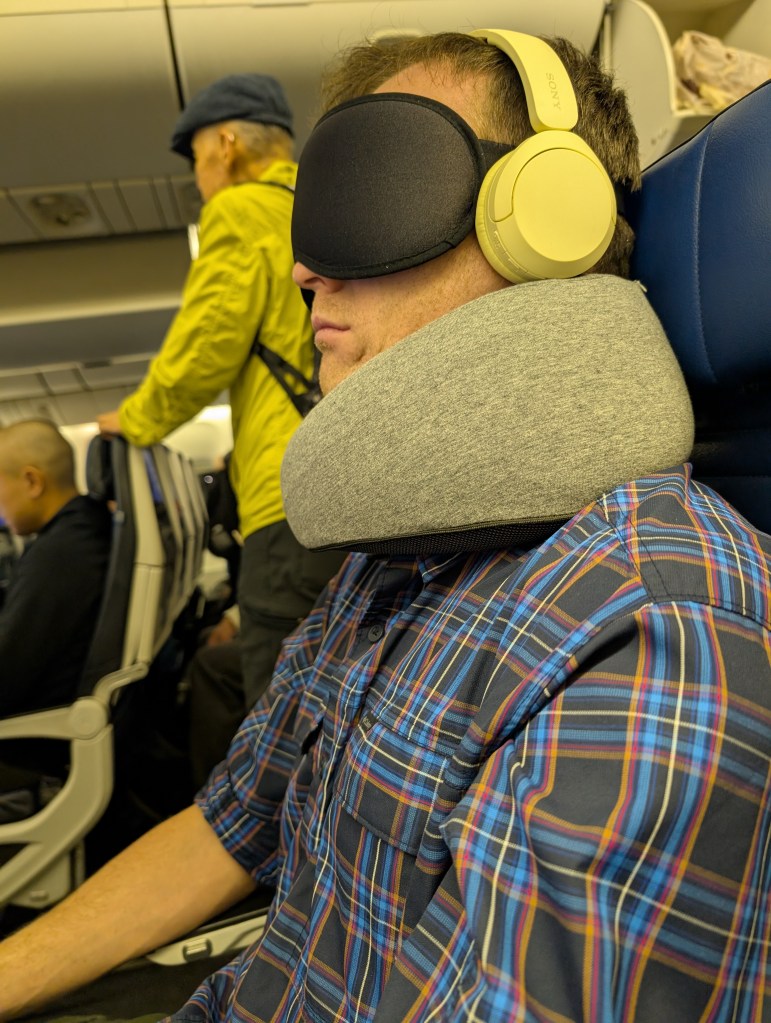 a man sits on plane with headphones, an eye mask, and a very thick neck pillow on.